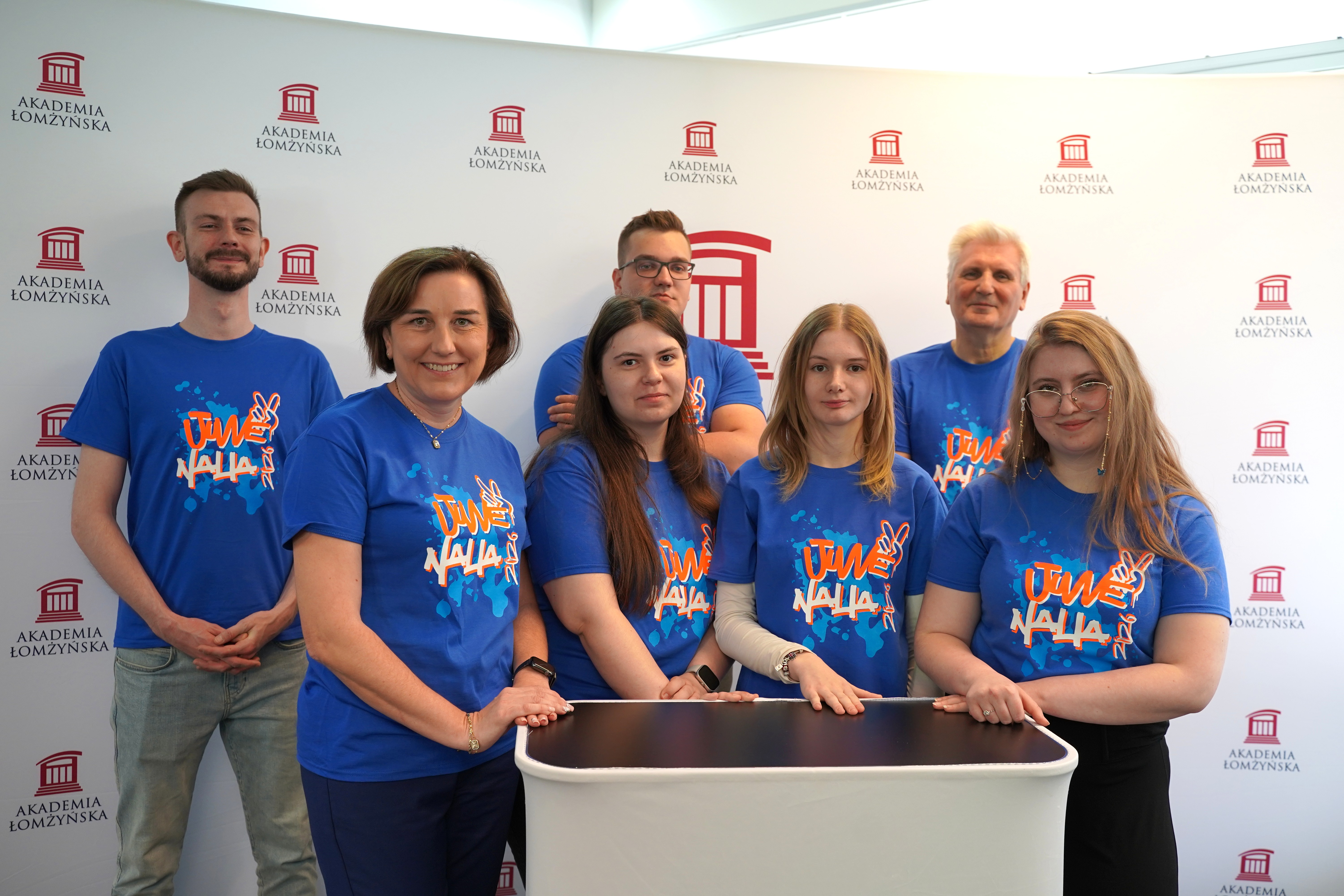 Grupa siedmiu osób w niebieskich koszulkach z nadrukiem „Juwenalia 2023” stoi i opiera ręce o mały, jasny stolik; w tle widoczna ścianka z powtarzającym się logo i napisem „Akademia Łomżyńska”