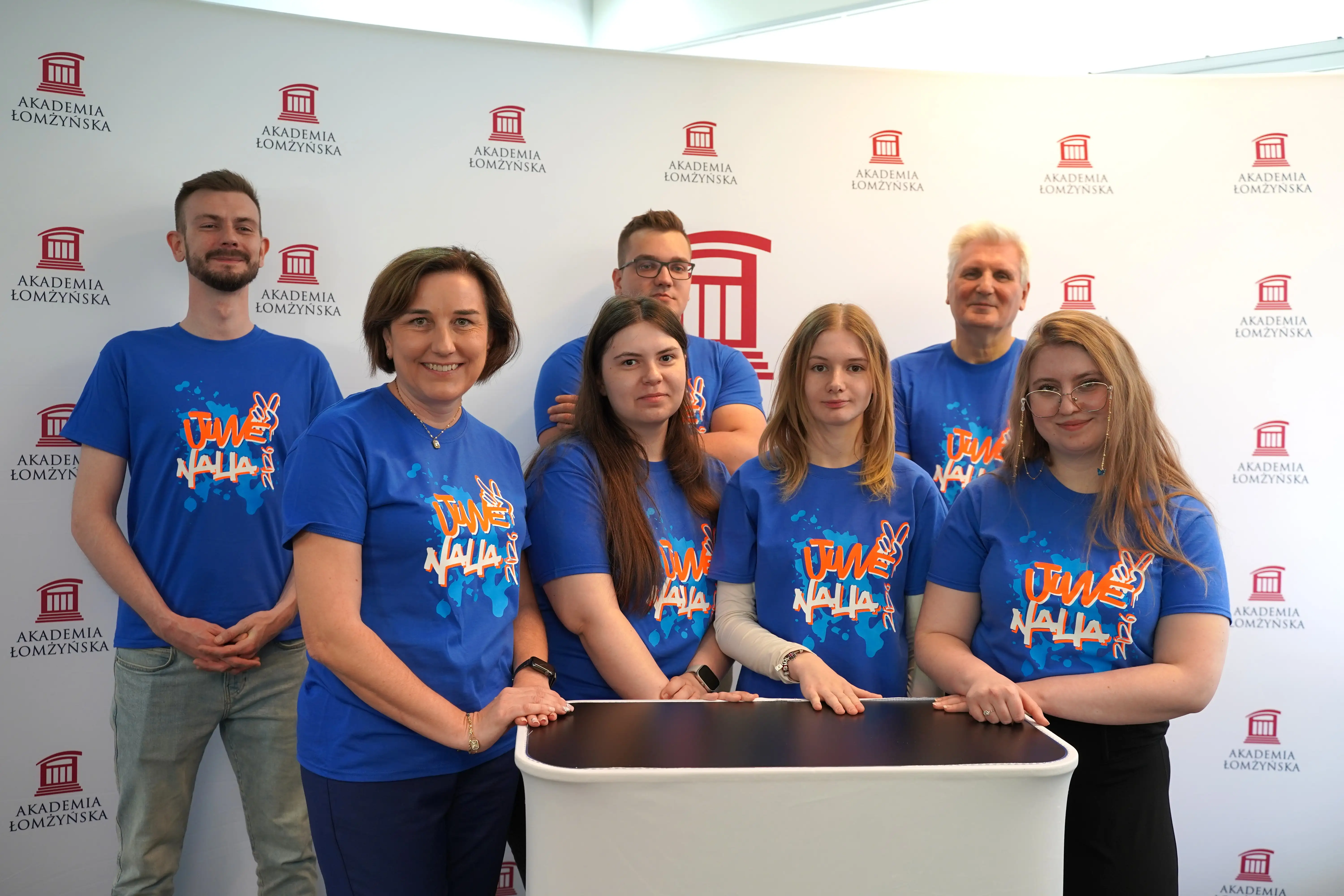 Grupa siedmiu osób w niebieskich koszulkach z nadrukiem „Juwenalia 2023” stoi i opiera ręce o mały, jasny stolik; w tle widoczna ścianka z powtarzającym się logo i napisem „Akademia Łomżyńska”