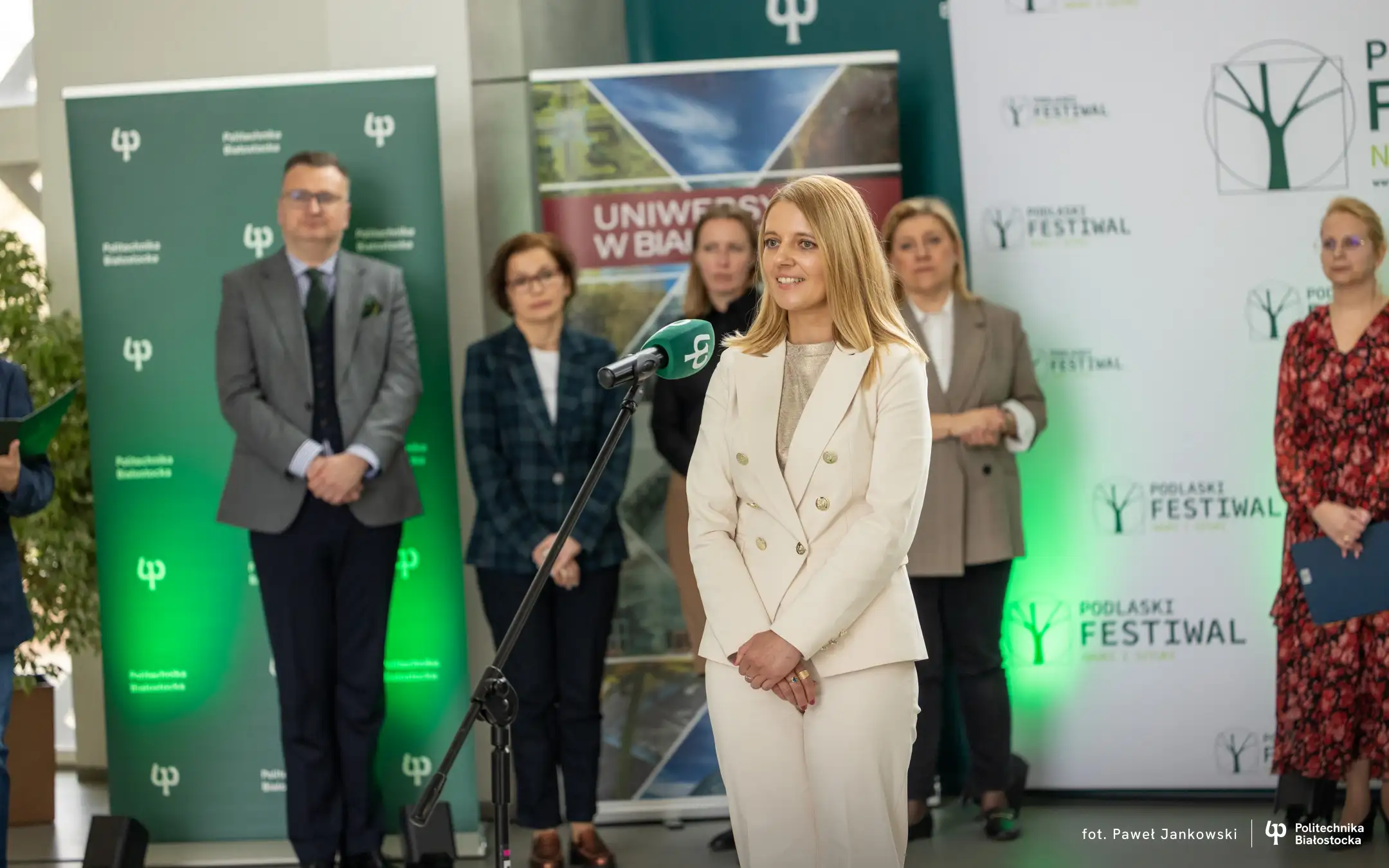 Osoba przemawiająca przy mikrofonie podczas wydarzenia naukowego; w tle stoją uczestnicy oraz banery Politechniki Białostockiej i Podlaskiego Festiwalu Nauki i Sztuki.