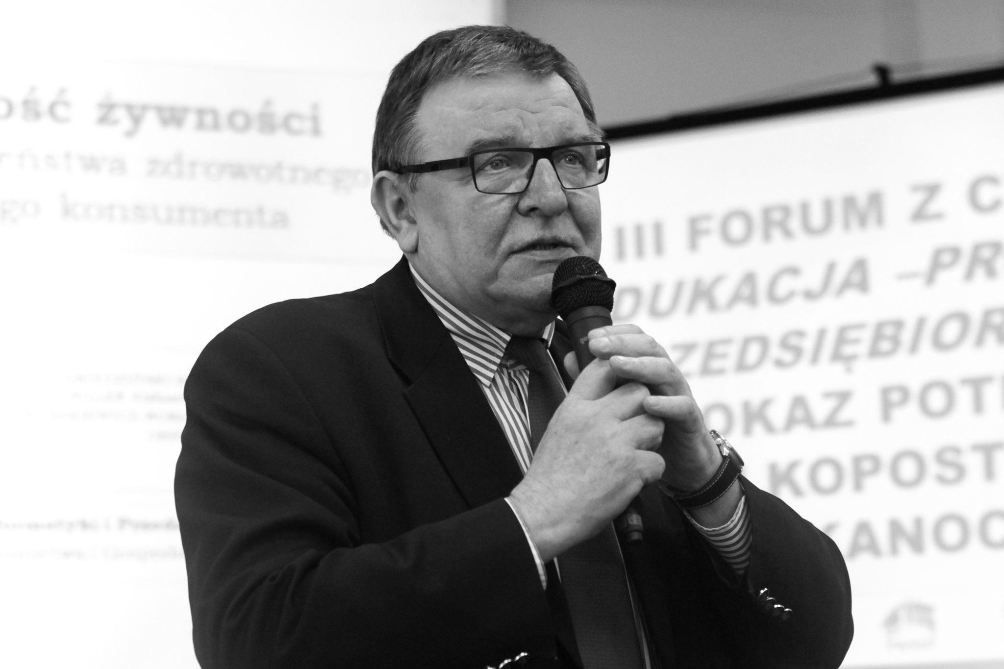 Czarno-biała fotografia osoby w garniturze trzymającej mikrofon i przemawiającej podczas konferencji, na tle slajdu prezentacyjnego