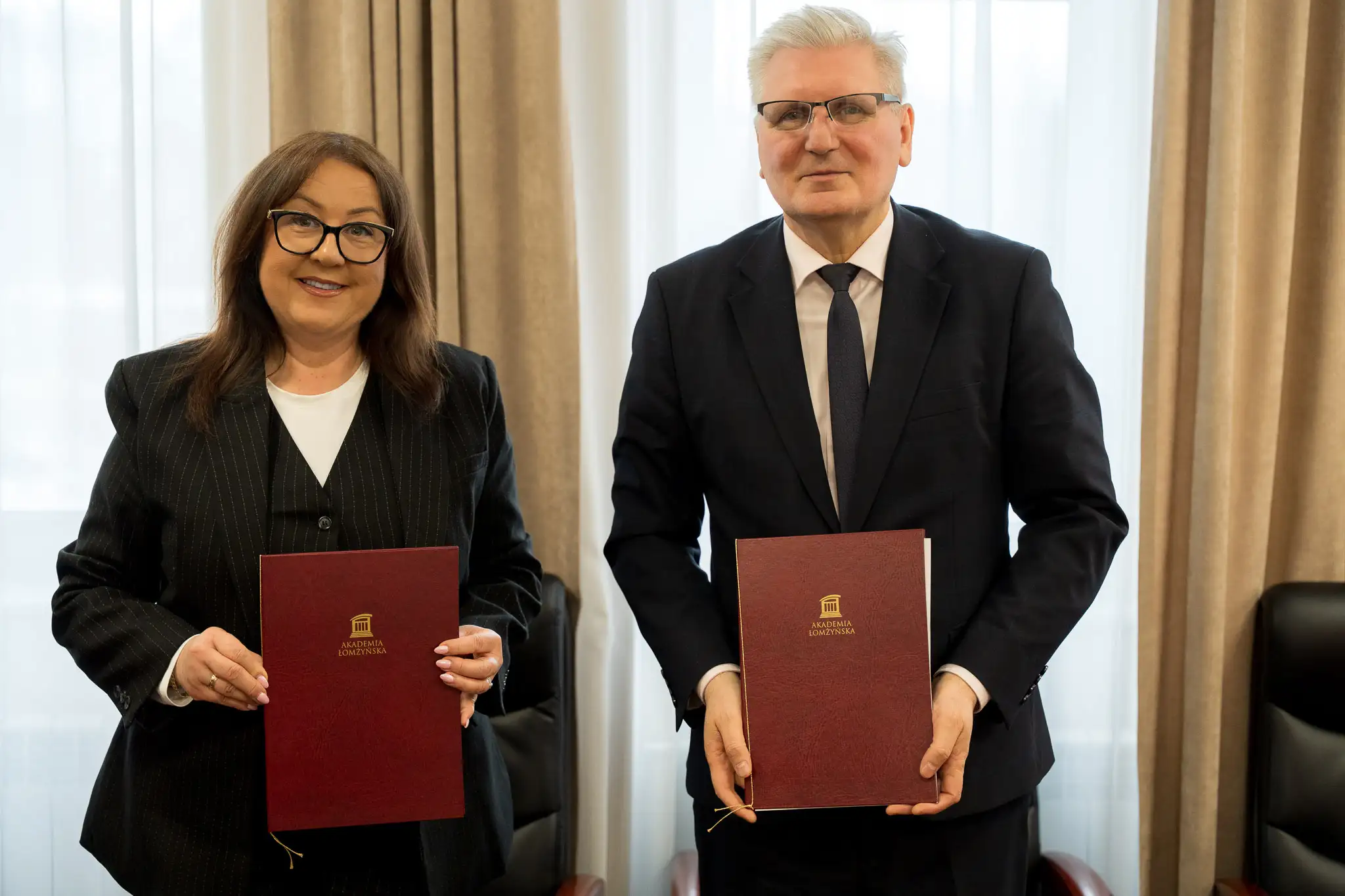 wie osoby stoją obok siebie w jasnym pomieszczeniu, każda trzyma bordową teczkę z widocznym złotym logo instytucji. Za nimi znajduje się duże okno z jasnymi zasłonami.