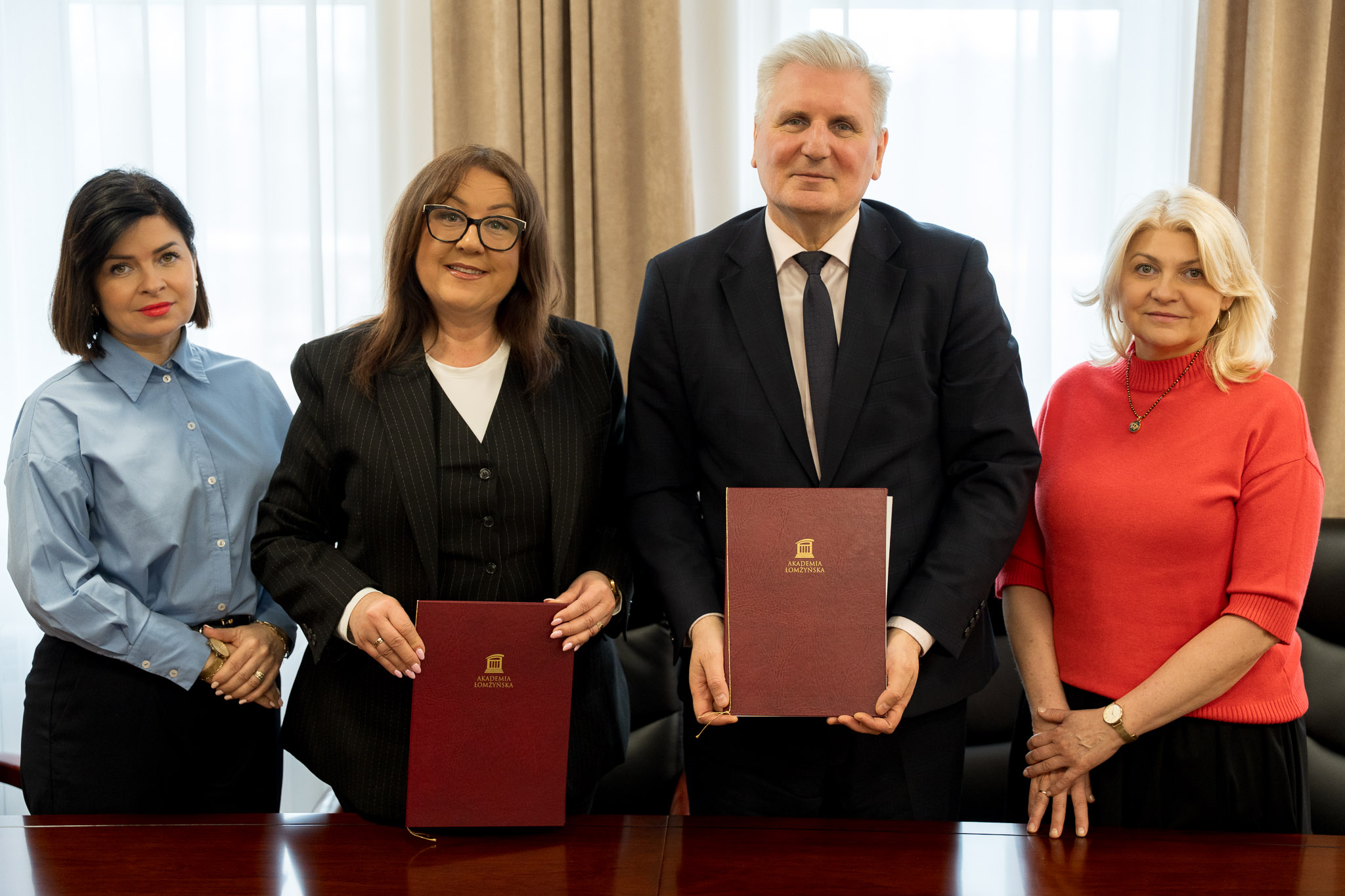 Cztery osoby stoją przy stole w jasnym pomieszczeniu, dwie z nich trzymają bordowe teczki z widocznym złotym logo instytucji. W tle znajdują się długie zasłony i duże okno