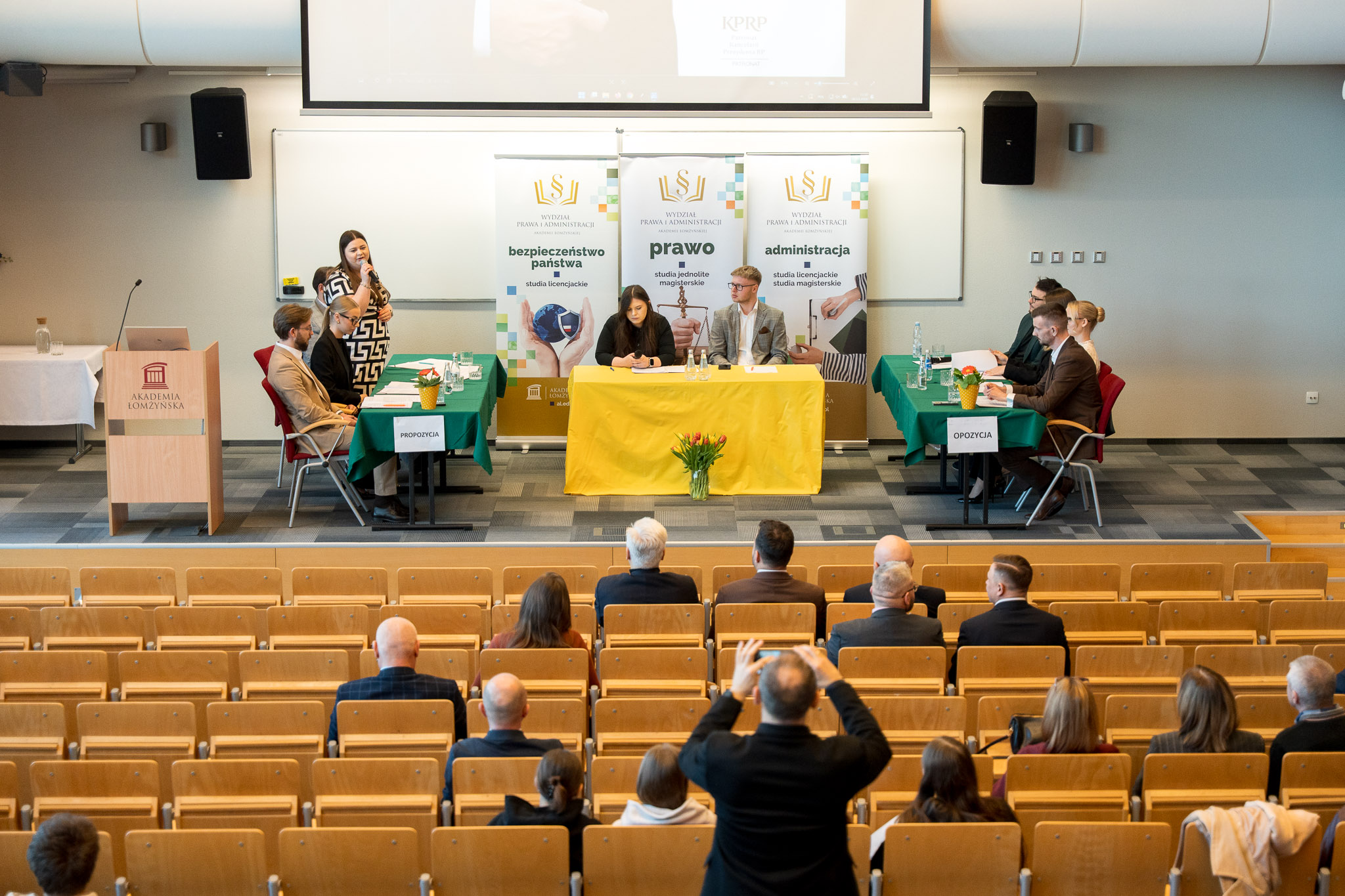 Zdjęcie przedstawia panel naukowy odbywający się w dużej sali wykładowej. Przy trzech stołach ustawionych w kształt litery U siedzą uczestnicy wydarzenia, a osoba stojąca z boku mówi do mikrofonu. Za centralnym stołem widoczne są trzy roll‑upy z informacjami o kierunkach: bezpieczeństwo państwa, prawo i administracja. Na widowni siedzi grupa osób obserwujących przebieg spotkania.