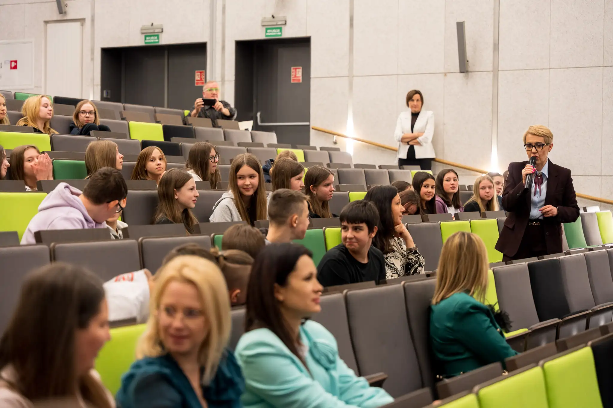 Wykład w dużej sali audytoryjnej, gdzie liczna grupa osób siedzi w rzędach, a jedna osoba stoi przy przejściu i przemawia do publiczności.