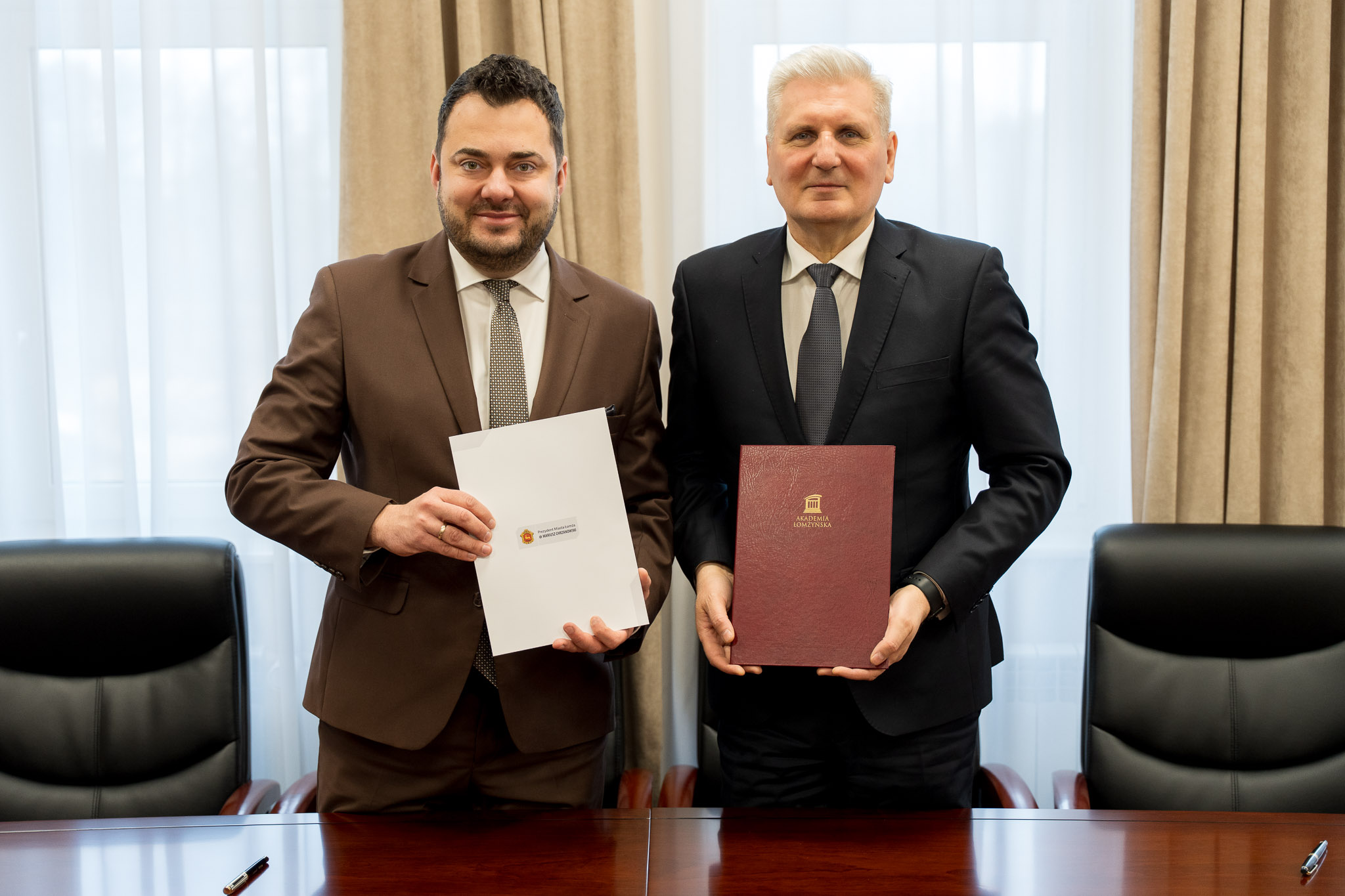 Dwie osoby w formalnych strojach prezentują podpisane dokumenty, stojąc przy stole w sali konferencyjnej.