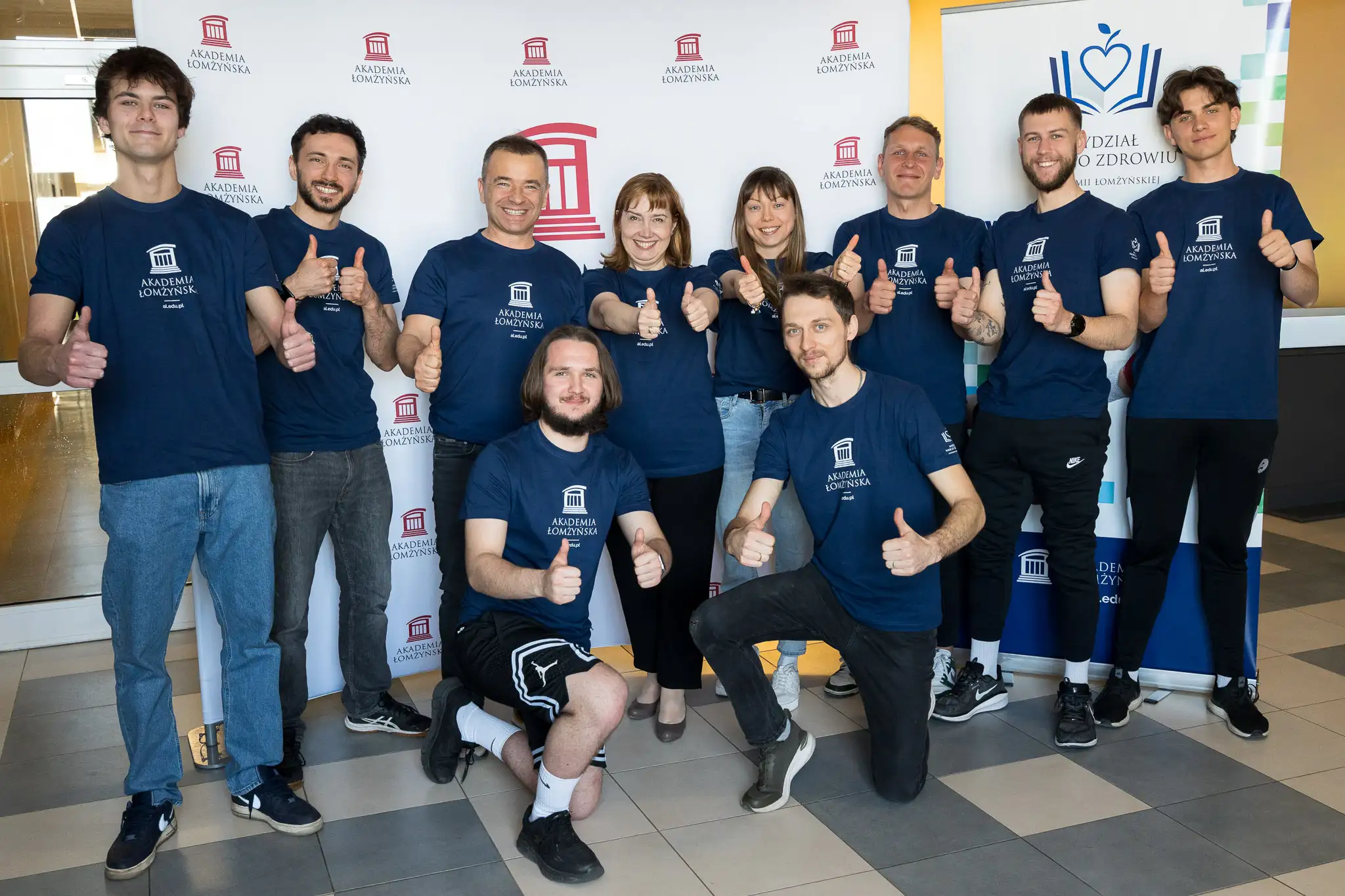 Grupa osób w koszulkach Akademii Łomżyńskiej pozuje do wspólnego zdjęcia przed ścianką z logo uczelni, wykonując gest podniesionego kciuka