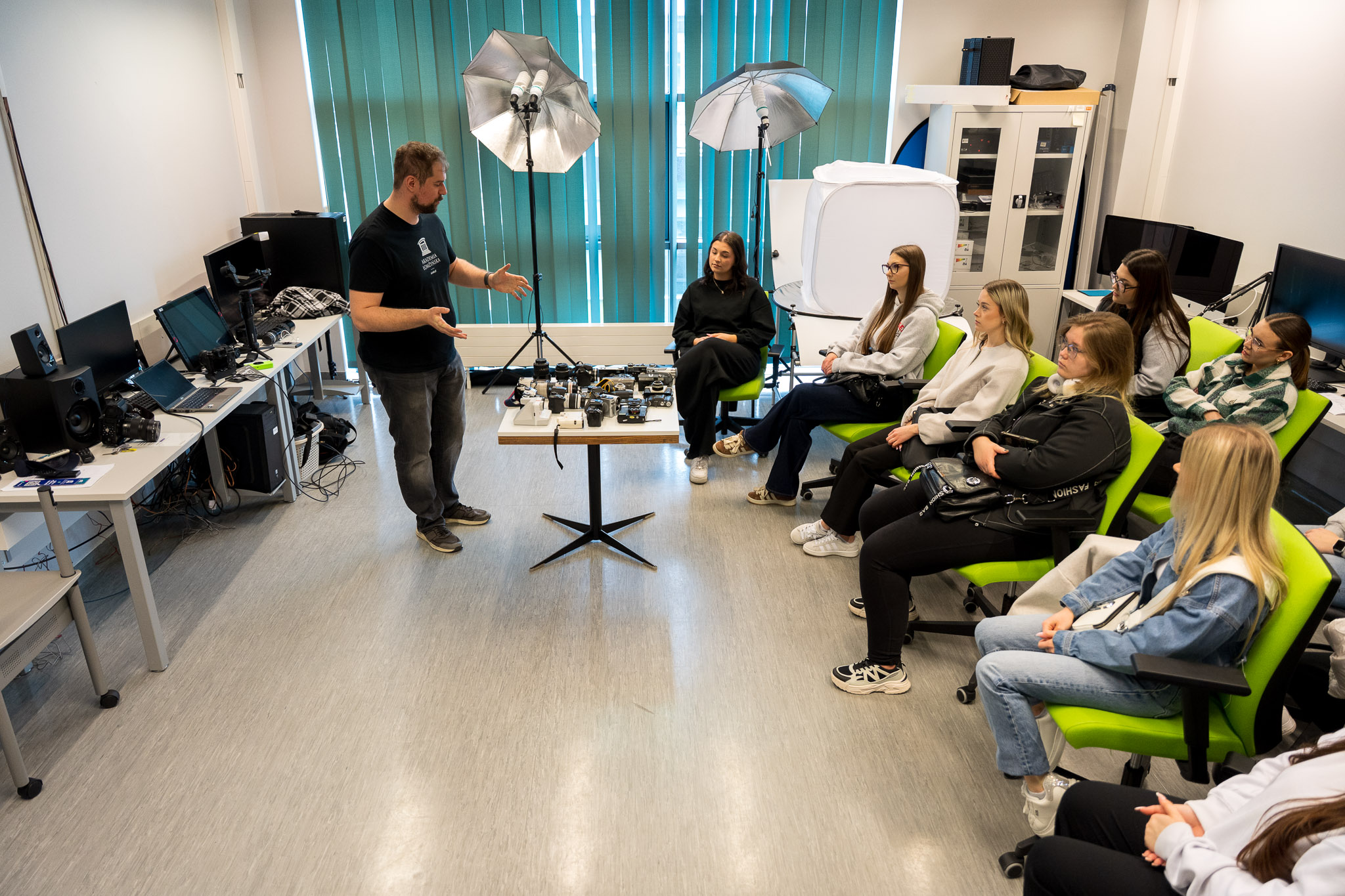Prowadzący prezentuje sprzęt fotograficzny i studyjne oświetlenie grupie uczestników siedzących w sali warsztatowej