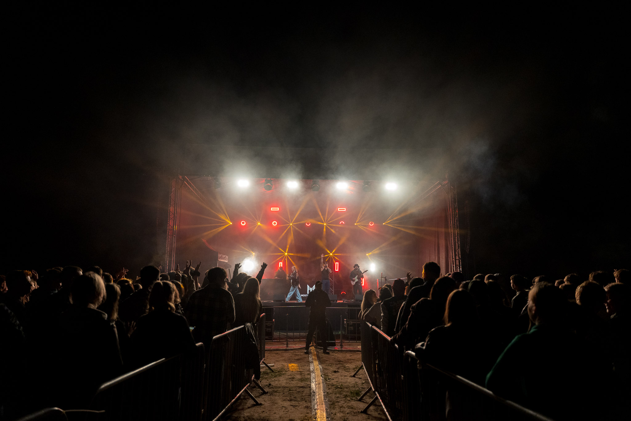 Koncert na otwartej scenie nocą, z zespołem oświetlonym kolorowymi reflektorami oraz publicznością stojącą przed sceną.