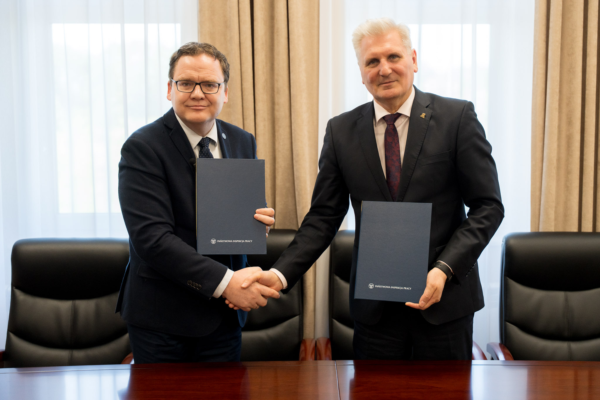 Dwie osoby w formalnych strojach stoją przy stole, podają sobie ręce i trzymają podpisane dokumenty podczas oficjalnego spotkania