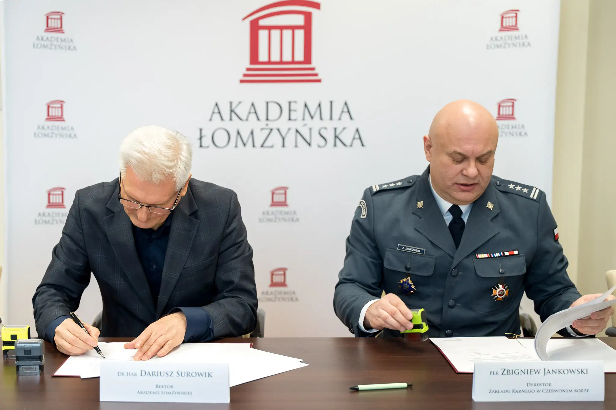 Dwóch mężczyzn siedzi przy stole i podpisuje dokumenty, na tle ścianki z logo Akademii Łomżyńskiej.