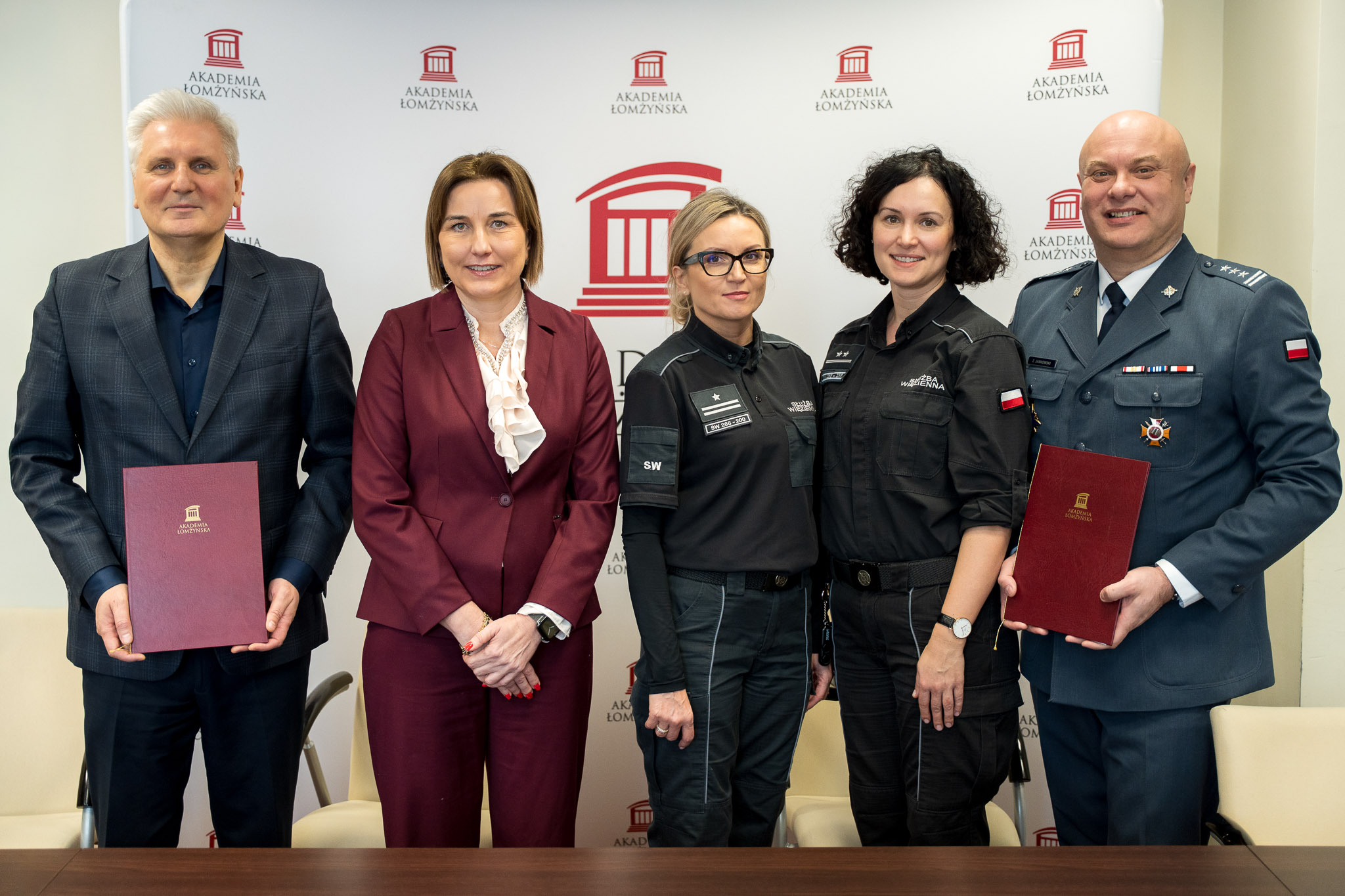 Grupa pięciu osób w strojach służbowych i formalnych stoi przed ścianką z logo Akademii Łomżyńskiej, trzymając teczki i pozując do wspólnego zdjęcia.