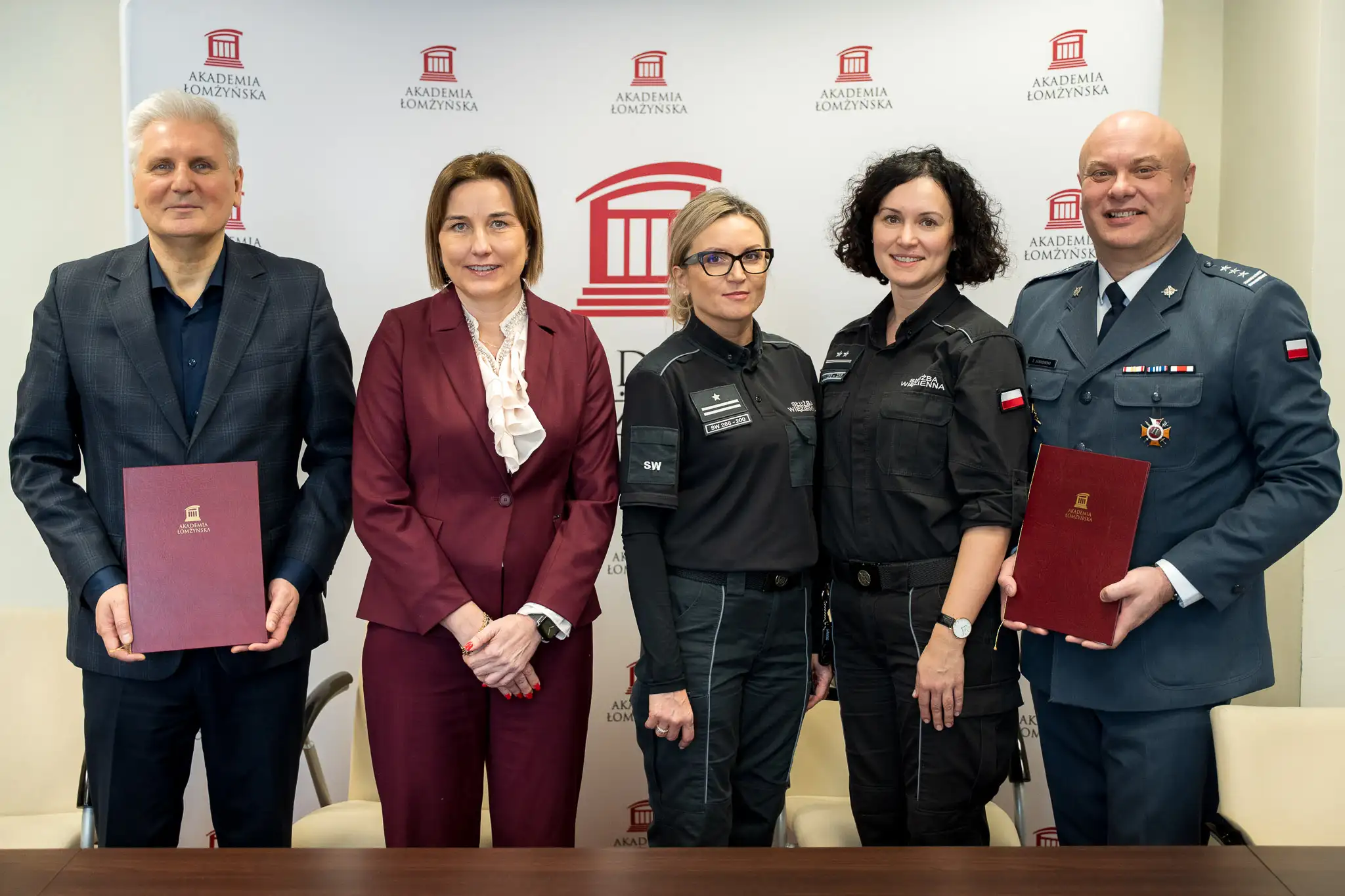 Grupa pięciu osób w strojach służbowych i formalnych stoi przed ścianką z logo Akademii Łomżyńskiej, trzymając teczki i pozując do wspólnego zdjęcia.