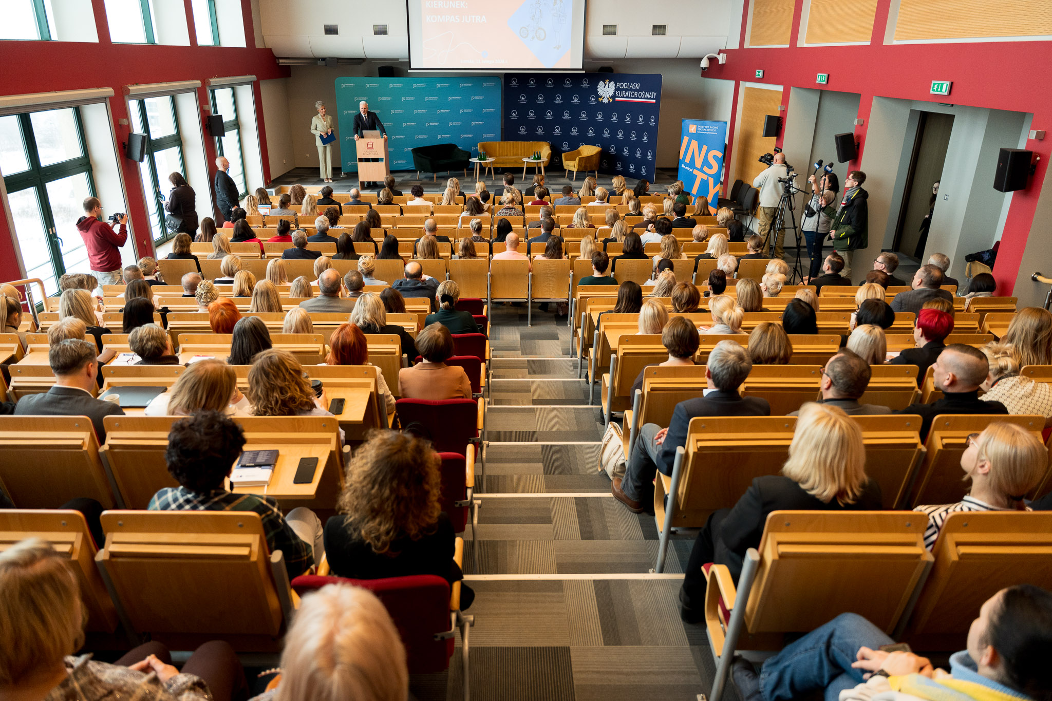 Duża sala wykładowa z licznie zgromadzonymi uczestnikami siedzącymi w rzędach, słuchającymi wystąpienia osoby stojącej przy mównicy na scenie.