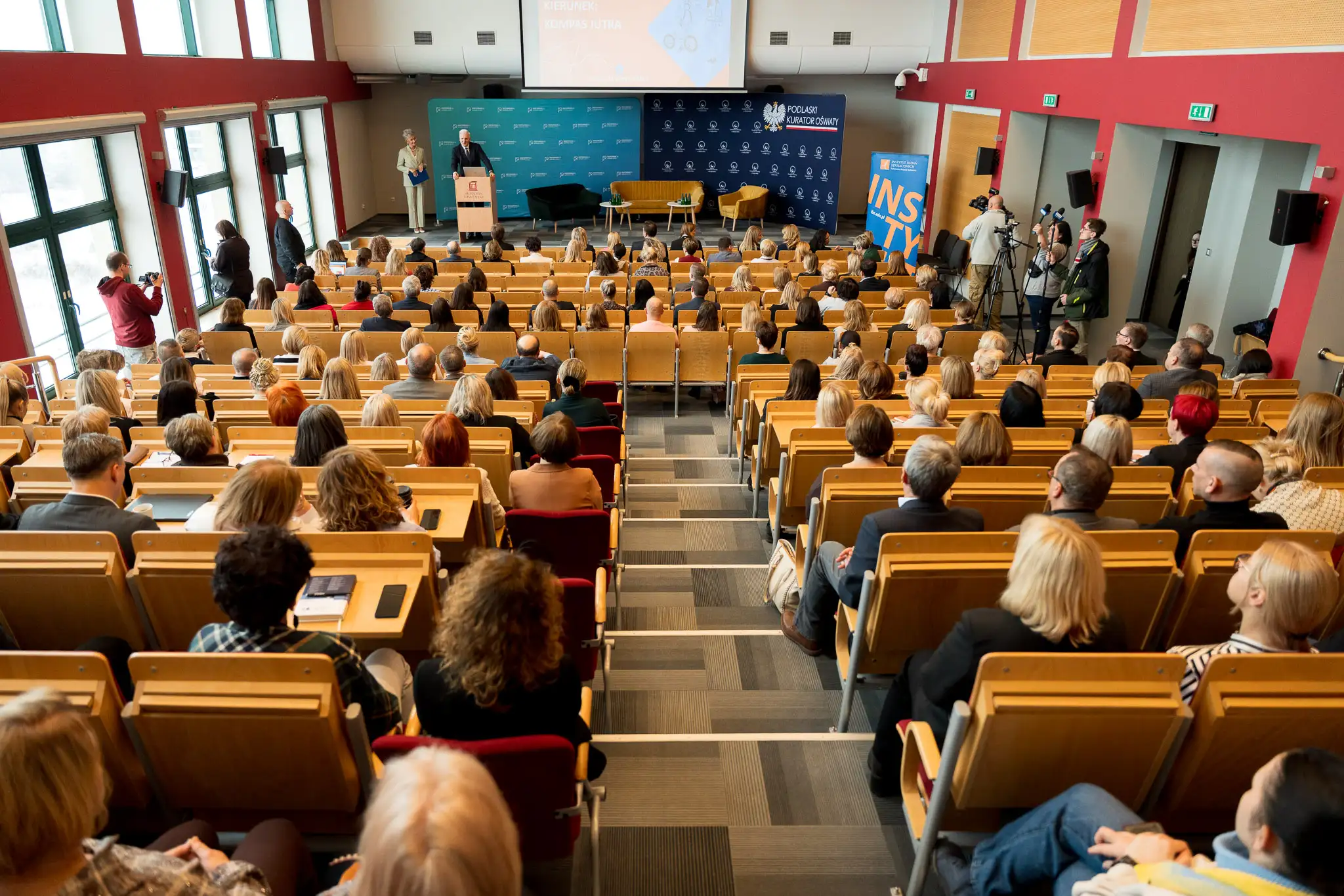 Duża sala wykładowa z licznie zgromadzonymi uczestnikami siedzącymi w rzędach, słuchającymi wystąpienia osoby stojącej przy mównicy na scenie.