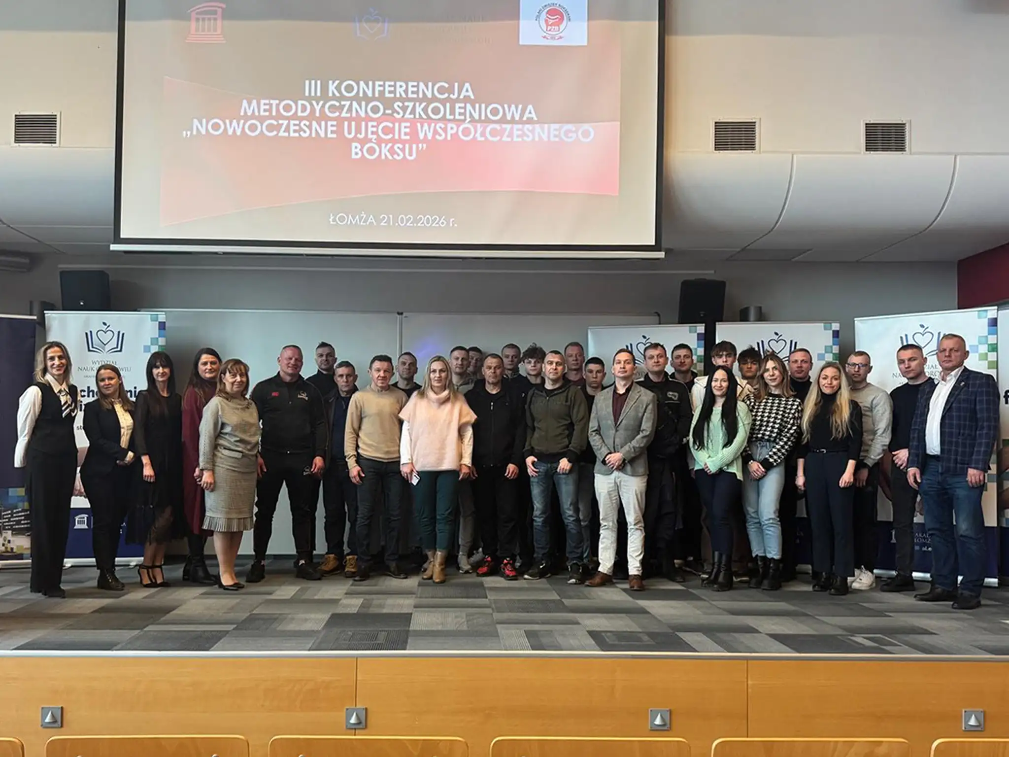 uża grupa uczestników stoi wspólnie na scenie w sali konferencyjnej przed ekranem wyświetlającym tytuł wydarzenia: ‘III Konferencja Metodyczno‑Szkoleniowa – Nowoczesne ujęcie współczesnego boksu’. Po obu stronach widoczne są roll‑upy organizatorów.