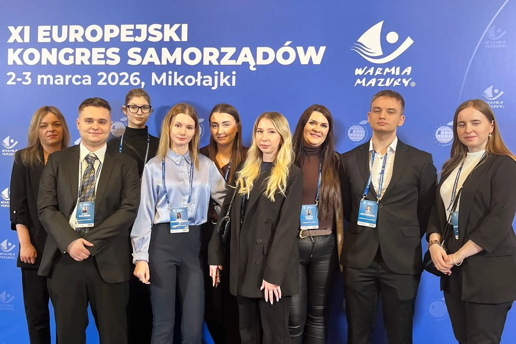 Grupa osób stoi przed niebieską ścianką konferencyjną z nadrukiem ‘XI Europejski Kongres Samorządów, 2–3 marca 2026, Mikołajki’ oraz logotypem Warmii i Mazur. Wszyscy mają identyfikatory zawieszone na smyczach.