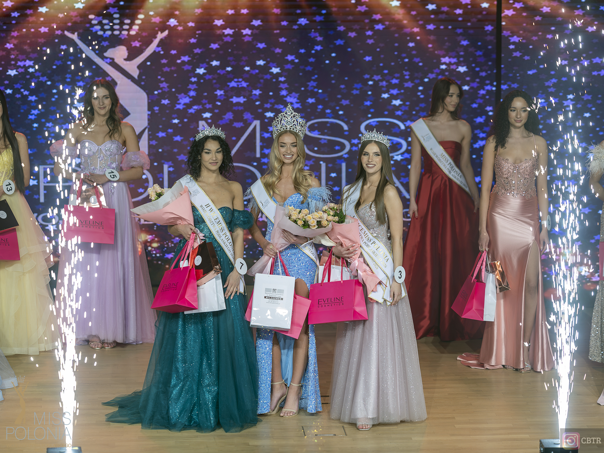 Uczestniczki finału konkursu piękności stoją na scenie w eleganckich sukniach, trzymając nagrody i kwiaty, w otoczeniu efektów pirotechnicznych.