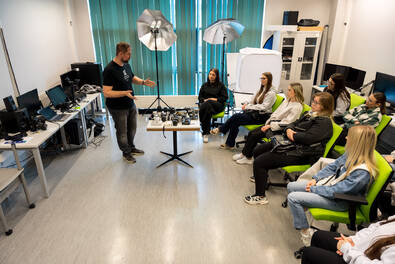 Prowadzący prezentuje sprzęt fotograficzny i studyjne oświetlenie grupie uczestników siedzących w sali warsztatowej