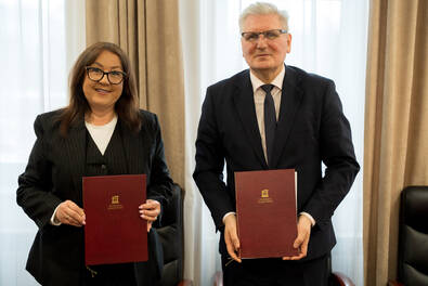 wie osoby stoją obok siebie w jasnym pomieszczeniu, każda trzyma bordową teczkę z widocznym złotym logo instytucji. Za nimi znajduje się duże okno z jasnymi zasłonami.