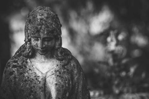 Czarno-biała fotografia kamiennej figury anioła z pochyloną głową, na rozmytym tle cmentarza