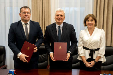 Trzy osoby stoją przy stole konferencyjnym, trzymając teczki z dokumentami w bordowych okładkach z widocznym złotym logo