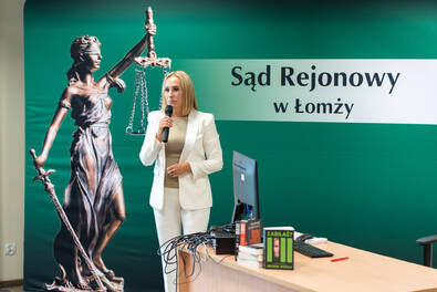 Osoba przemawia do mikrofonu podczas spotkania w sali, na tle grafiki z Temidą i napisem ‘Sąd Rejonowy w Łomży