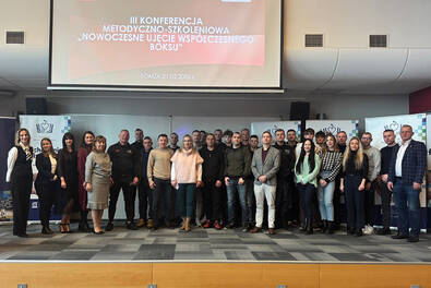 uża grupa uczestników stoi wspólnie na scenie w sali konferencyjnej przed ekranem wyświetlającym tytuł wydarzenia: ‘III Konferencja Metodyczno‑Szkoleniowa – Nowoczesne ujęcie współczesnego boksu’. Po obu stronach widoczne są roll‑upy organizatorów.