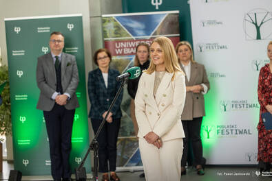 Osoba przemawiająca przy mikrofonie podczas wydarzenia naukowego; w tle stoją uczestnicy oraz banery Politechniki Białostockiej i Podlaskiego Festiwalu Nauki i Sztuki.