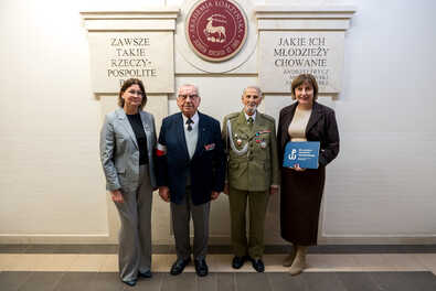 Cztery osoby stoją przed tablicą z napisem Akademia Łomżyńska, w tym dwie w strojach wojskowych, jedna trzyma niebieską teczkę.