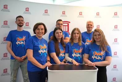 Grupa siedmiu osób w niebieskich koszulkach z nadrukiem „Juwenalia 2023” stoi i opiera ręce o mały, jasny stolik; w tle widoczna ścianka z powtarzającym się logo i napisem „Akademia Łomżyńska”