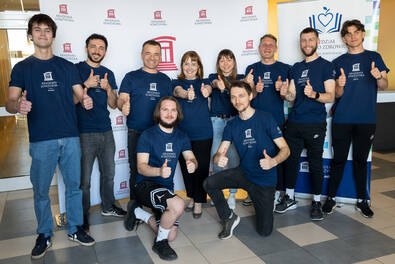 Grupa osób w koszulkach Akademii Łomżyńskiej pozuje do wspólnego zdjęcia przed ścianką z logo uczelni, wykonując gest podniesionego kciuka