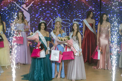 Uczestniczki finału konkursu piękności stoją na scenie w eleganckich sukniach, trzymając nagrody i kwiaty, w otoczeniu efektów pirotechnicznych.