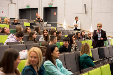 Wykład w dużej sali audytoryjnej, gdzie liczna grupa osób siedzi w rzędach, a jedna osoba stoi przy przejściu i przemawia do publiczności.