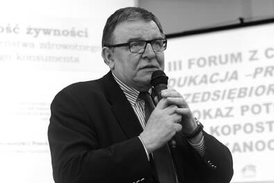 Czarno-biała fotografia osoby w garniturze trzymającej mikrofon i przemawiającej podczas konferencji, na tle slajdu prezentacyjnego