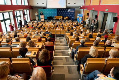 Duża sala wykładowa z licznie zgromadzonymi uczestnikami siedzącymi w rzędach, słuchającymi wystąpienia osoby stojącej przy mównicy na scenie.