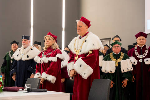 Osoby ubrane w uroczyste stroje akademickie stoją podczas ceremonii w sali konferencyjnej, przy stole prezydialnym