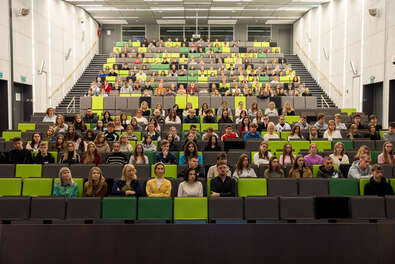Duża aula wykładowa z wieloma rzędami zielono-szarych siedzeń, wypełniona uczestnikami wydarzenia.