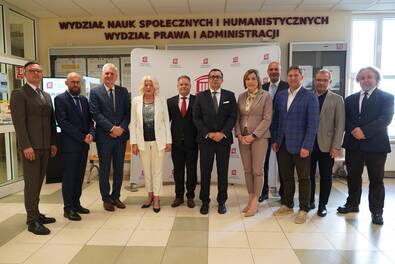 Grupa jedenastu osób w formalnych strojach stoi do wspólnego zdjęcia w holu Akademii Łomżyńskiej, na tle ścianki z logo uczelni oraz napisu „Wydział Nauk Społecznych i Humanistycznych, Wydział Prawa i Administracji