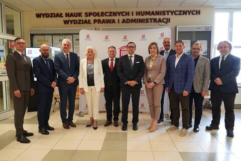 Grupa jedenastu osób w formalnych strojach stoi do wspólnego zdjęcia w holu Akademii Łomżyńskiej, na tle ścianki z logo uczelni oraz napisu „Wydział Nauk Społecznych i Humanistycznych, Wydział Prawa i Administracji