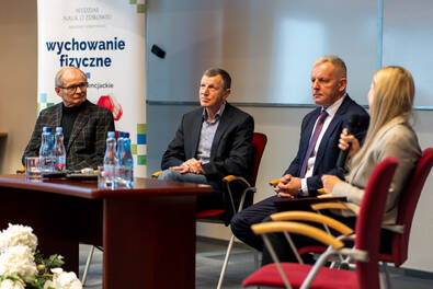 Cztery osoby siedzą na krzesłach w sali konferencyjnej podczas panelu dyskusyjnego. Trzej mężczyźni w garniturach i kobieta z mikrofonem biorą udział w rozmowie. Na stole przed nimi stoją butelki z wodą. W tle widoczny jest baner z napisem „Wydział Nauk o Zdrowiu – Wychowanie fizyczne” oraz biała tablica.