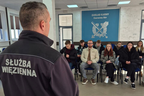 Mężczyzna w kurtce z napisem „Służba Więzienna” stoi tyłem do kamery i rozmawia z grupą młodych osób siedzących na krzesłach w sali. W tle widoczna jest niebieska ściana z napisem „Zakład Karny Czerwony Bór” oraz symbolem instytucji.