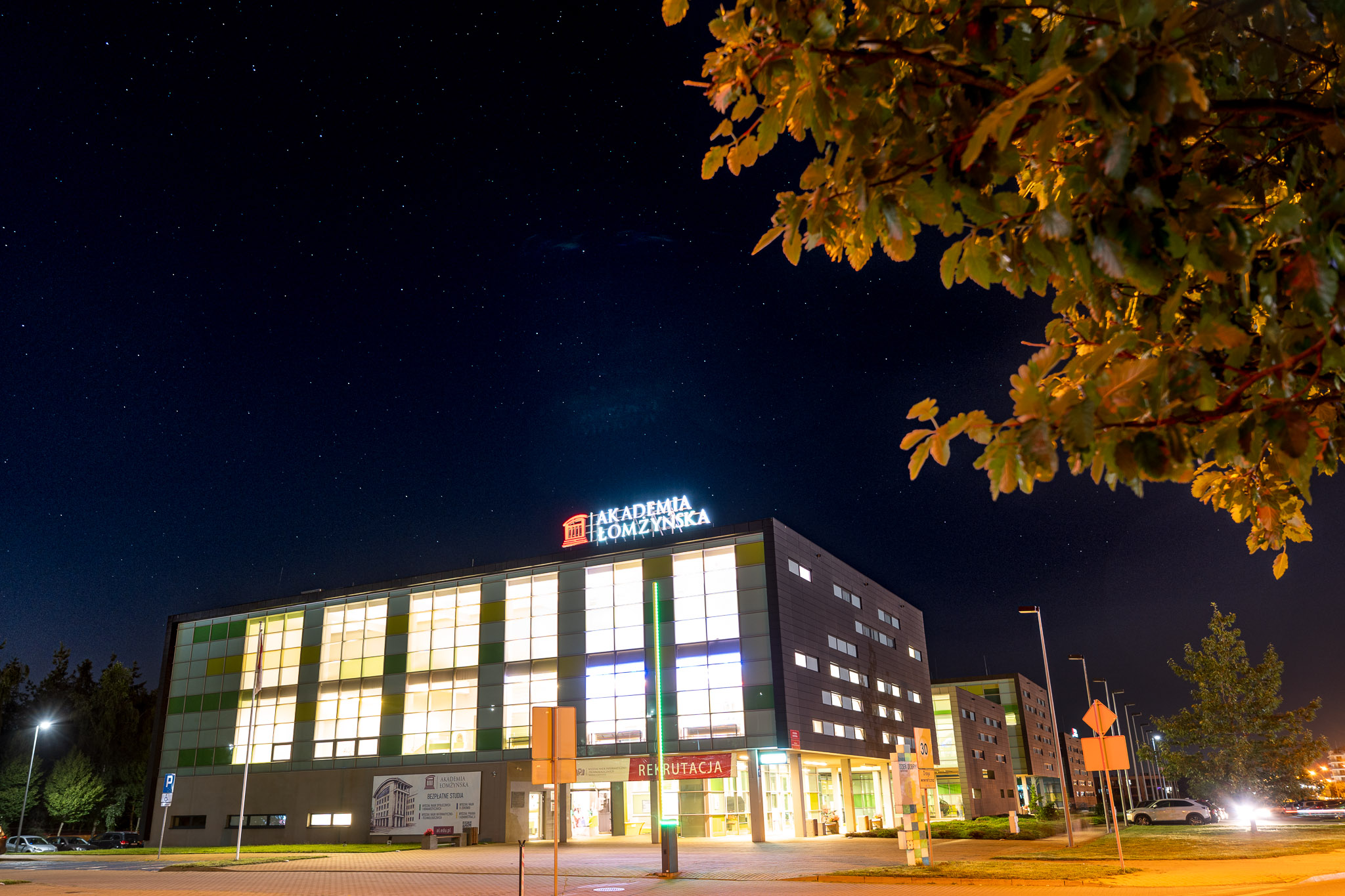 Modern building of Akademia Łomżyńska at night under a starry sky, illuminated by streetlights and surrounded by trees.