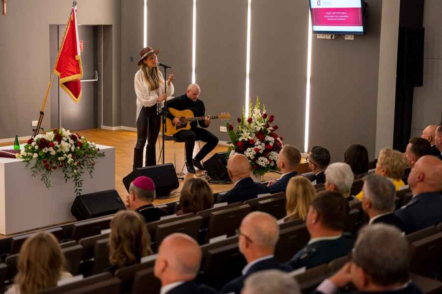 Na scenie stoi kobieta w brązowym kapeluszu, śpiewa do mikrofonu. Obok niej na krześle siedzi gitarzysta.
