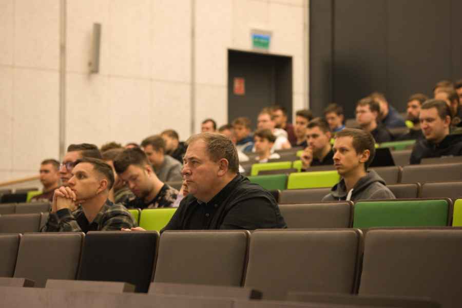 Wnętrze dużej sali wykładowej z rzędami nowoczesnych foteli w odcieniach zieleni i szarości. Na miejscach siedzi grupa osób, skierowanych w stronę przodu sali, co sugeruje trwający wykład lub prezentację.