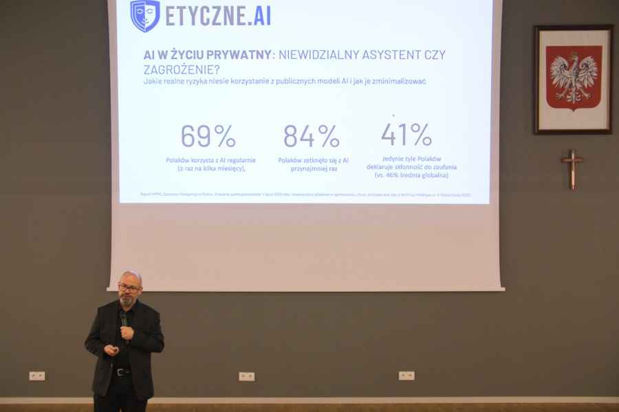 Osoba stoi przed dużym ekranem w sali konferencyjnej lub wykładowej, prezentując slajd 