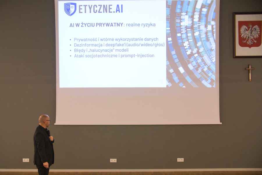 Osoba stoi przed dużym ekranem w sali konferencyjnej lub wykładowej, prezentując slajd 