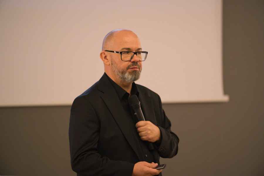 Osoba w ciemnym garniturze stoi przed jasnym ekranem w sali konferencyjnej, trzymając w jednej ręce mikrofon, a w drugiej pilot do prezentacji. 