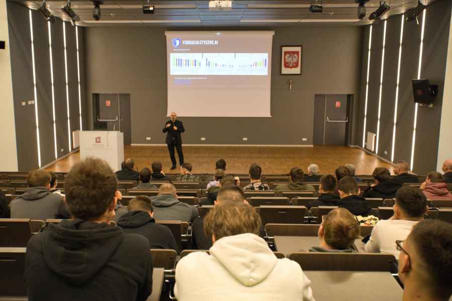 Widok z tyłu sali wykładowej w nowoczesnym budynku. Na pierwszym planie widać rzędy siedzących osób, skierowanych w stronę sceny. Na scenie stoi prelegent z mikrofonem, obok mównicy, przed dużym ekranem projekcyjnym. 