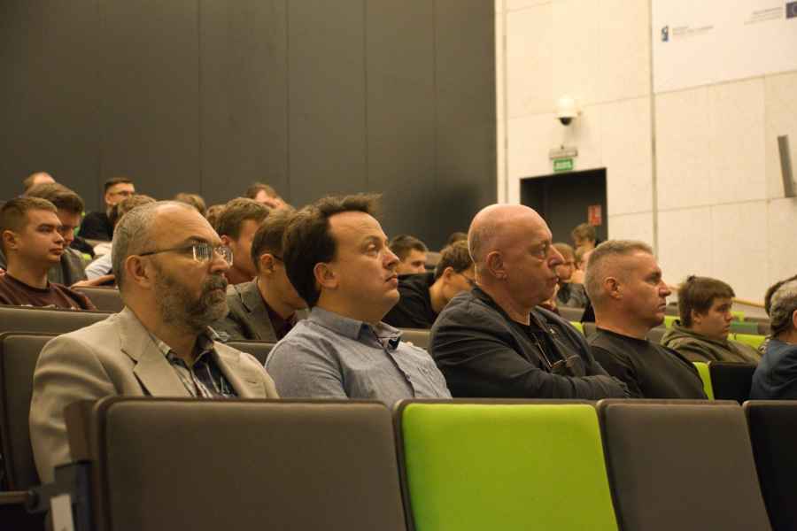 Wnętrze sali wykładowej z rzędami nowoczesnych foteli w odcieniach zieleni i szarości. Na miejscach siedzi grupa osób, skierowanych w stronę przodu sali, co sugeruje trwający wykład lub prezentację.