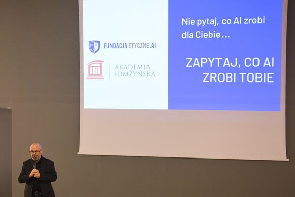 Osoba prowadząca wykład mówi przez mikrofon, za nią wyświetlana jest prezentacja, na której jest napisane "Nie pytaj co AI zrobi dla Ciebie...zapytaj co zrobi Tobie"